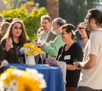 a group of people talking while standing around a blue table with yellow flowers in a white vase.